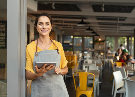 Portrait of happy woman standing at doorway of her franchise store holding digital tablet. Successful franchise business owner in casual clothing and grey apron standing at entrance and looking at camera.
